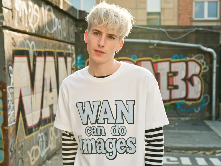 A cinematic, photorealistic medium shot capturing the nostalgic warmth of a mid-2000s indie film. The focus is a young man with a messy mullet haircut in cool platinum white, looking directly and intently into the camera lens with a knowing smirk, his head is looking up slightly. He wears an oversized band t-shirt that says “WAN can do Images" in huge stylized text over a long-sleeved striped top and simple silver stud earrings. The lighting is soft, golden hour sunlight creating lens flare and illuminating dust motes in the air. The background shows a blurred outdoor urban setting with graffiti-covered walls (the graffiti says “Wan" in stylized graffiti lettering), rendered with a shallow depth of field. Natural film grain, a warm, slightly muted color palette, and sharp focus on his expressive eyes enhance the intimate, authentic feel