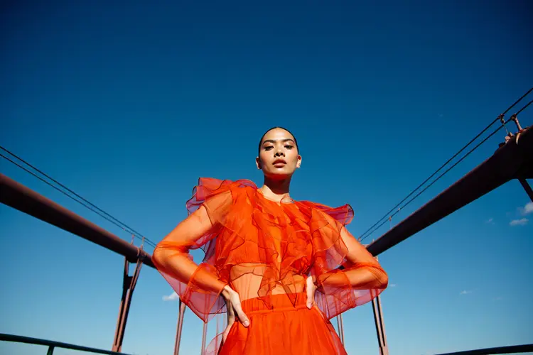 A powerful low-angle fashion portrait of a woman standing confidently in front of a large suspension bridge, shot on a bright, clear day. She wears a dramatic, avant-garde sheer orange ruffled outfit that catches the sunlight and adds rich texture and movement to the image. Her expression is poised and bold, with the shot capturing her from below to emphasize power and presence. The bridge's architectural lines frame her dynamically against a deep blue sky with minimal clouds. The image is captured in 8K resolution with a cinematic color grade — high contrast, slightly warm highlights, crisp shadows, and a soft filmic filter that enhances the drama and editorial quality of the scene. The overall mood is empowering, high-fashion, and editorial-worthy.