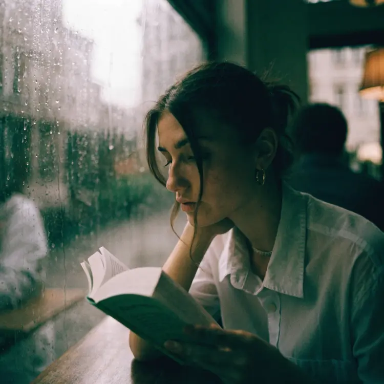 A woman in her late 20s sitting by a rain-streaked window in a dim café, holding a paperback book, soft ambient light, candid and unposed, 35mm film aesthetic, shallow depth of field, photorealistic.