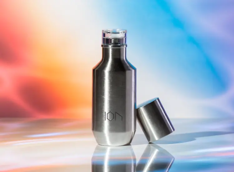 A playful commercial still life of a sleek metallic minimalist perfume bottle. Brushed steel or matte aluminum finish, clean geometric silhouette, cap casually placed beside the bottle. Engraved on the front: subtle "ION" monogram, minimal and refined, precisely etched into the metal surface (no other logos or text). The engraving catches the light softly, creating elegant micro-reflections. Set on a smooth pastel surface. Bright vibrant gradient background (peach to coral or sky blue to lilac). Glossy reflections, soft diffused studio lighting, crisp highlights and gentle shadows. Add subtle motion light streaks or color reflections for energy. Modern commercial fragrance aesthetic — fun, stylish, energetic, premium yet approachable.